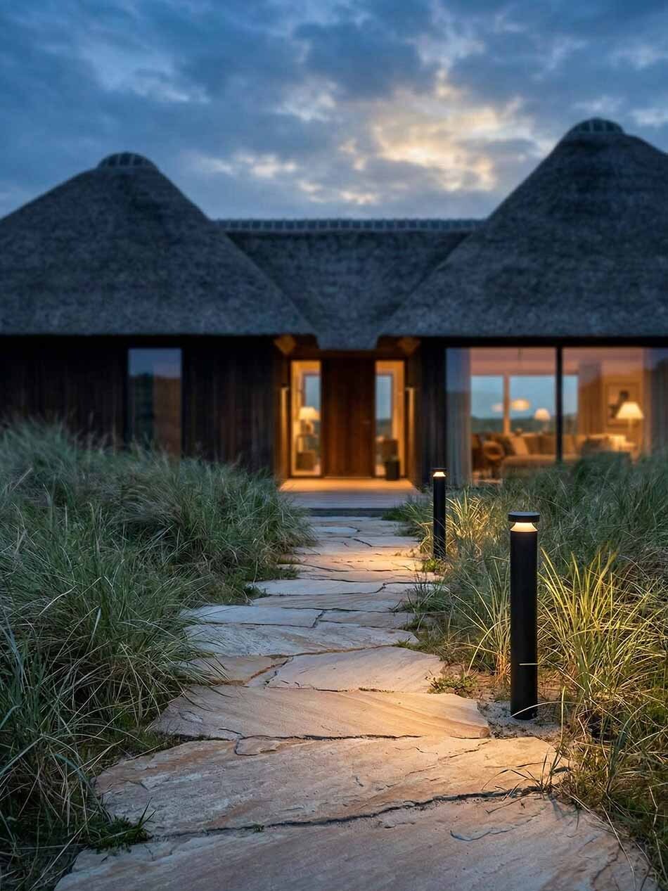 Moderne schwarze Pollerleuchte p.146 von prediger.base entlang eines Naturstein-Gartenwegs bei Dämmerung. Die schlanke LED-Außenleuchte spendet warmes, blendfreies Rundumlicht zwischen hohen Gräsern vor einem modernen Haus mit Reetdach.