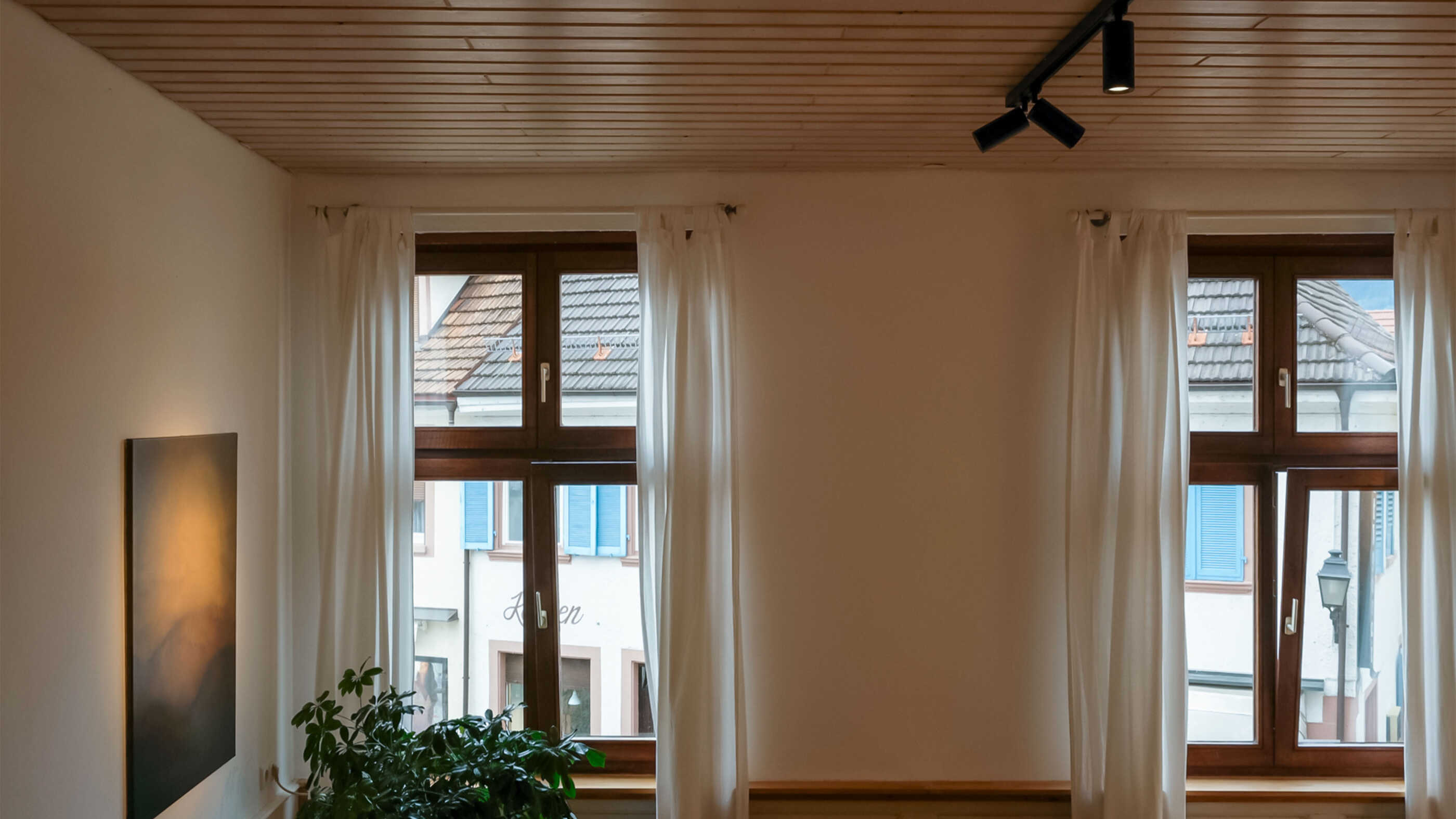 Drei Schienenstrahler an einer Schiene beleuchten ein Wohnzimmer. Gezielt wird das Bild auf der linken Seite angestrahlt, zwei Fenster (voraus) sorgen für Tageslicht.
