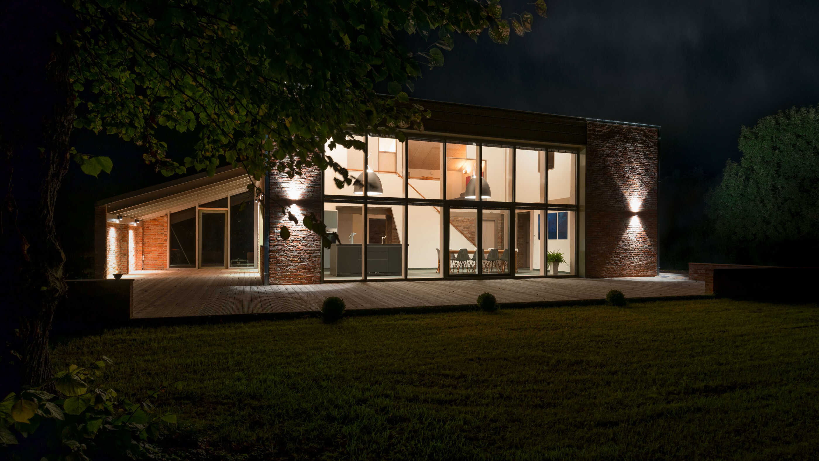 Ein Ferienhaus in Schleswig-Holstein bei Nacht aus leicht schräger Perspektive: Die zweigeschossige Fensterfront erstreckt sich fast über die gesamte Breite des Hauses und ist hell erleuchtet, Wandleuchten links und rechts rahmen das Fenster ein. (Foto: Prediger Lichtberater)