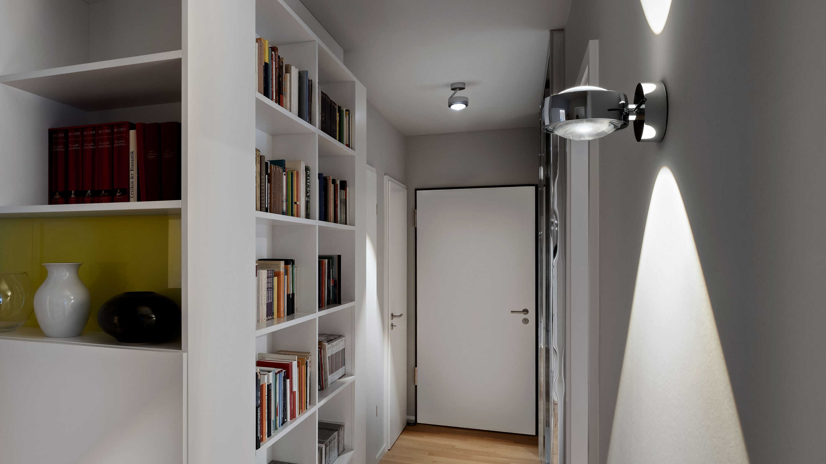 Ein Flur wird auf der rechten Seite von Wandleuchten von Occhio beleuchtet. Links steht ein wandhohes Bücherregal. Ein Deckenstrahler betont die Wohnungs-Eingangstür. (Foto: Prediger Lichtberater)