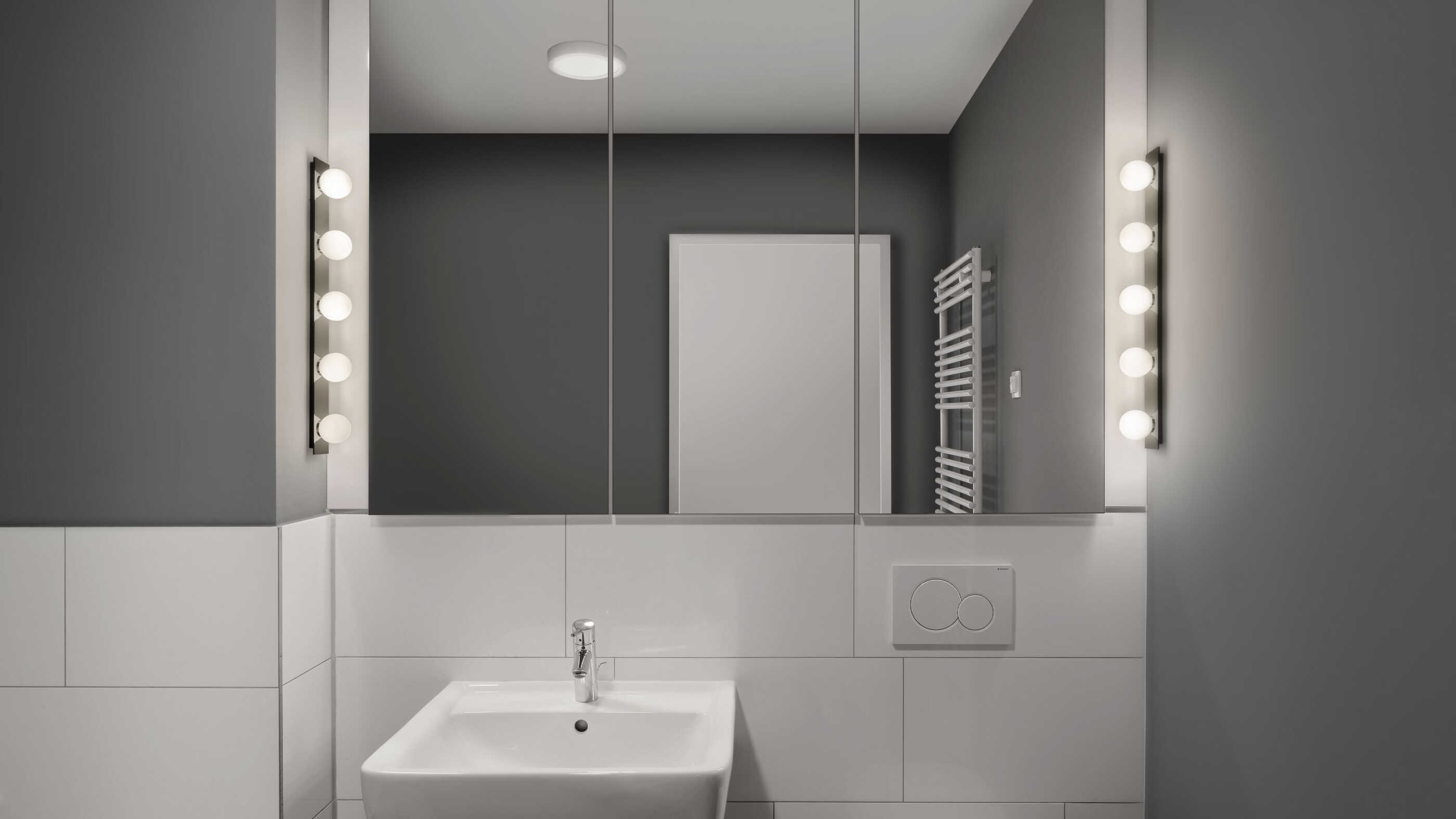 Ein minimalistisch eingerichtetes Badezimmer: Zwei Wandleuchten auf beiden Seiten des Spiegels sorgen für ausreichende Beleuchtung, das Raumlicht wird von einer Deckenleuchte in der Raummitte ergänzt. (Foto: Prediger Lichtberater)