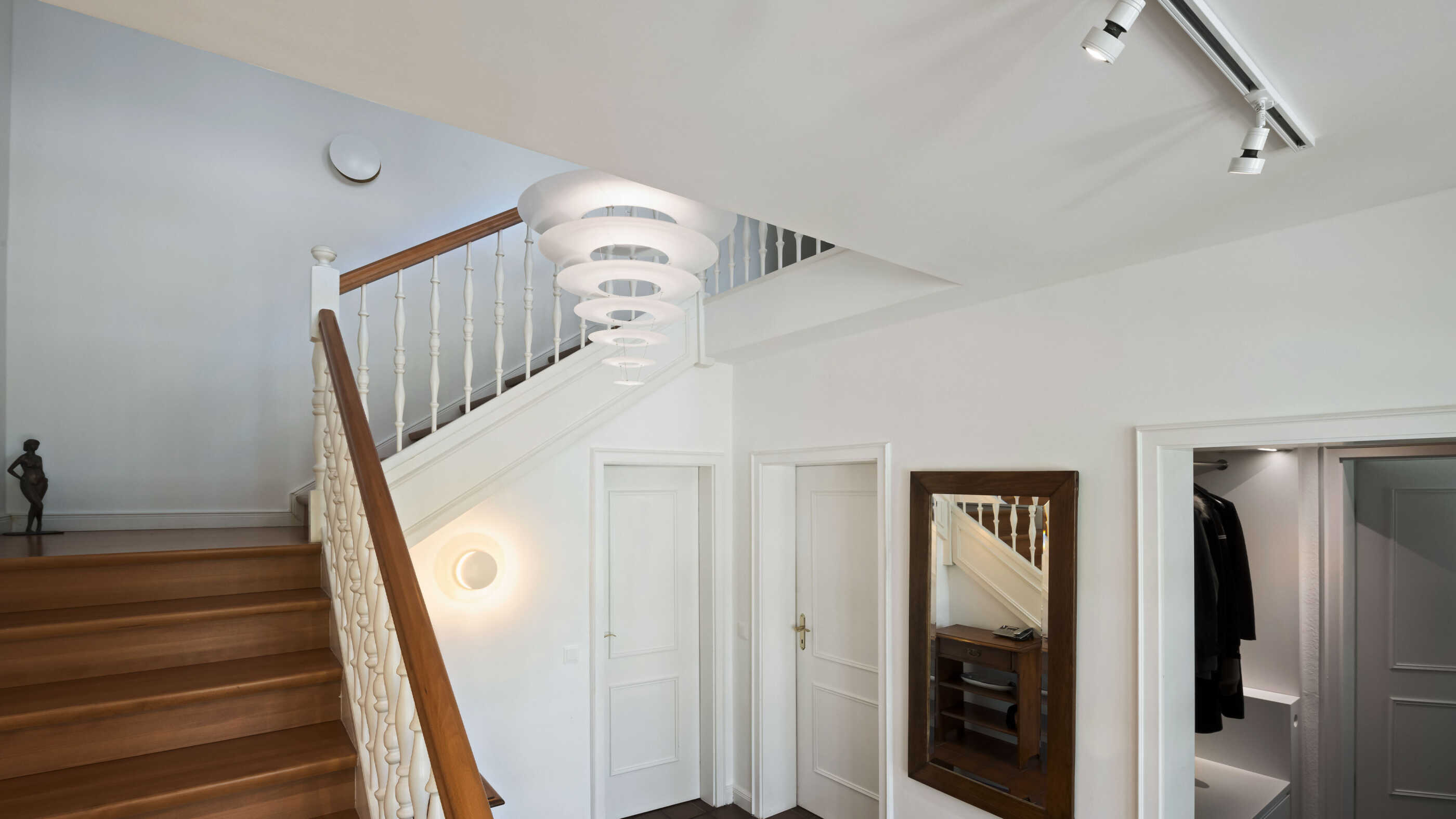 Ein stilvoll beleuchtetes Treppenhaus im Landhausstil, schräge Perspektive: Die Form der Pendelleuchte im Treppenauge erinnert an einen Kegel, der auf der Spitze steht. (Foto: Prediger Lichtberater)