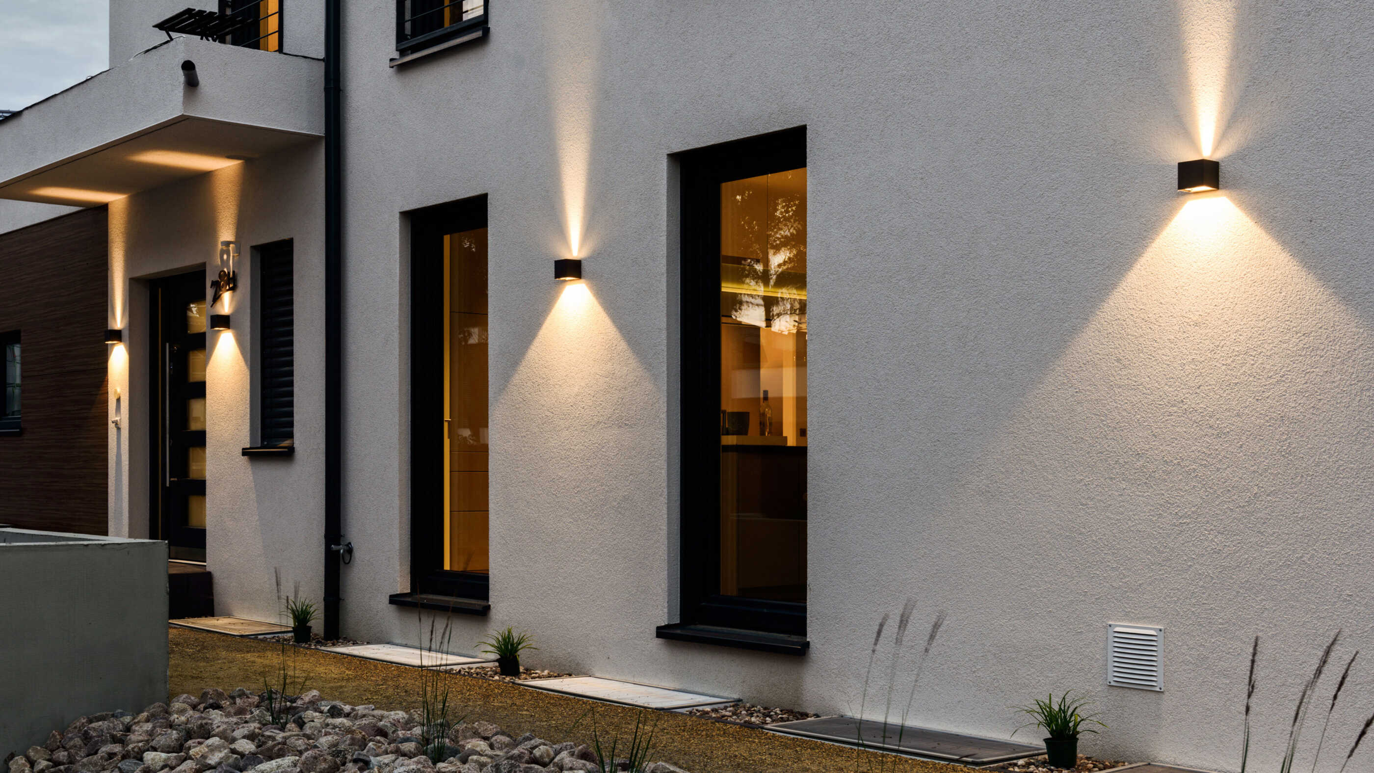 Eine Hausfassade wird von zweiseitig strahlenden Wandleuchten beleuchtet. Links im Bild ist die Haustür zu sehen, die von zwei Wandleuchten eingerahmt wird. (Foto: Prediger Lichtberater)