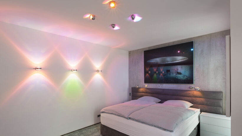 Das Schlafzimmer eines stylischen Penthouses in Hamburg: Drei Wandleuchten sowie vier Deckenleuchten mit Farbfiltern sorgen für eine ausgewogene und stimmungsvolle Grundbeleuchtung. (Foto: Prediger Lichtberater)