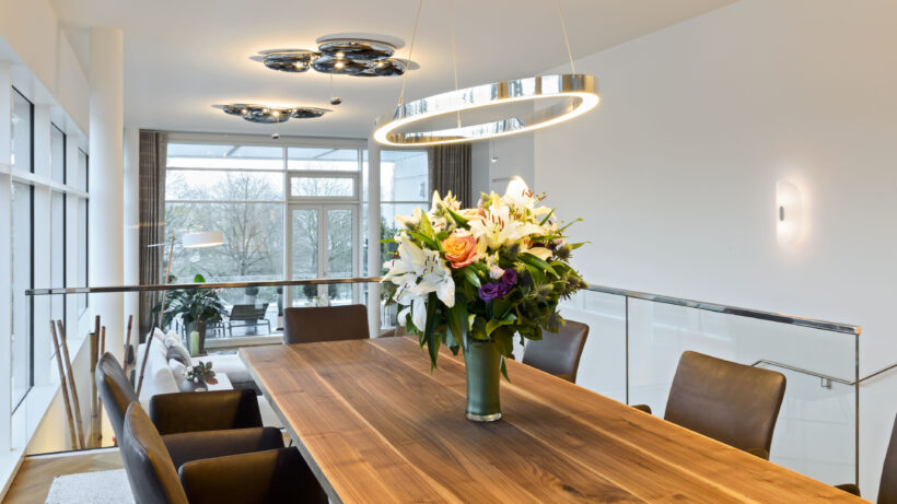 Das Esszimmer in einem modernen Penthouse an der Alster in Hamburg: Eine ovale Pendelleuchte über dem Blumenstrauß in der Mitte des Tisches sorgt für künstliches Licht, im Hintergrund sind die indirekt strahlenden Deckenleuchten des Wohnzimmers zu sehen. (Foto: Prediger Lichtberater)