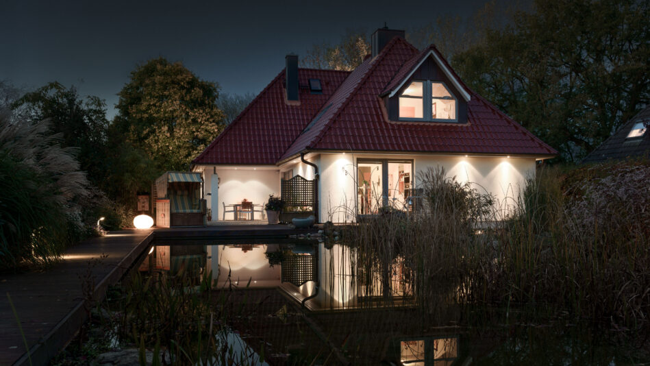 Ein stimmungsvoll beleuchtetes Haus bei Nacht: Die von Downlights beleuchtete Fassade spiegelt sich im benachbarten Gartenteich. (Foto: Prediger Lichtberater)