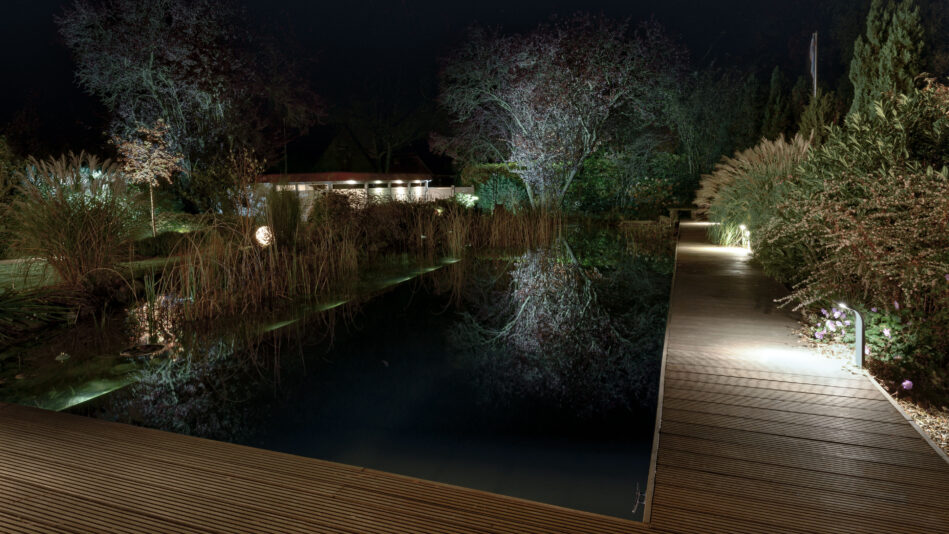 Ein Schwimmteich bei Nacht. Pollerleuchten an der Seite beleuchten den Rand des Pools. (Foto: Prediger Lichtberater)