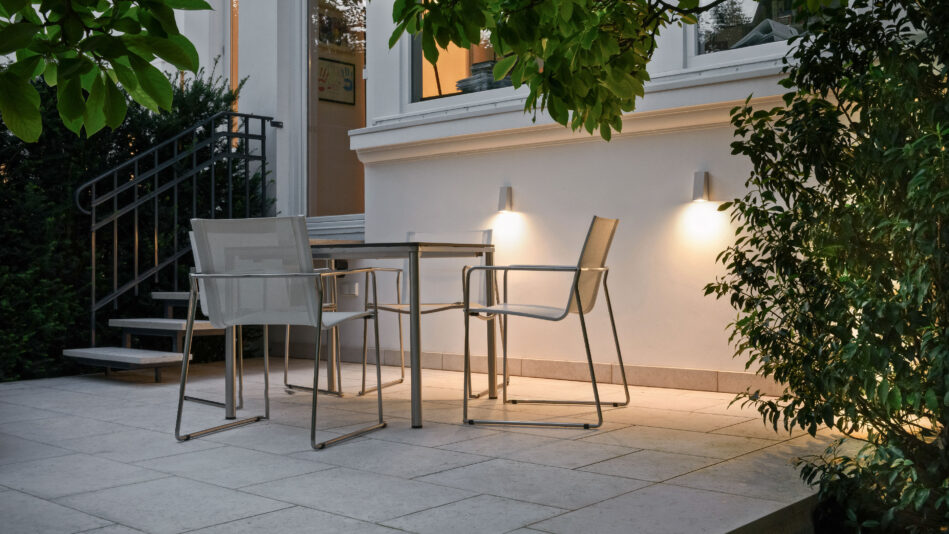 Ein Tisch und vier Stühle stehen auf einer Terrasse, die Wand dahinter wird von zwei Wandleuchten beleuchtet. (Foto: Prediger Lichtberater)