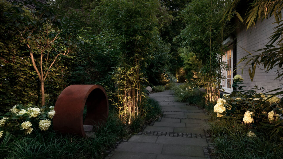 Ein gepflasterter Weg schlängelt sich durch einen üppig bewachsenen Garten, Spießstrahler in der Vegetation setzen dramatische Lichtakzente. (Foto: Prediger Lichtberater)