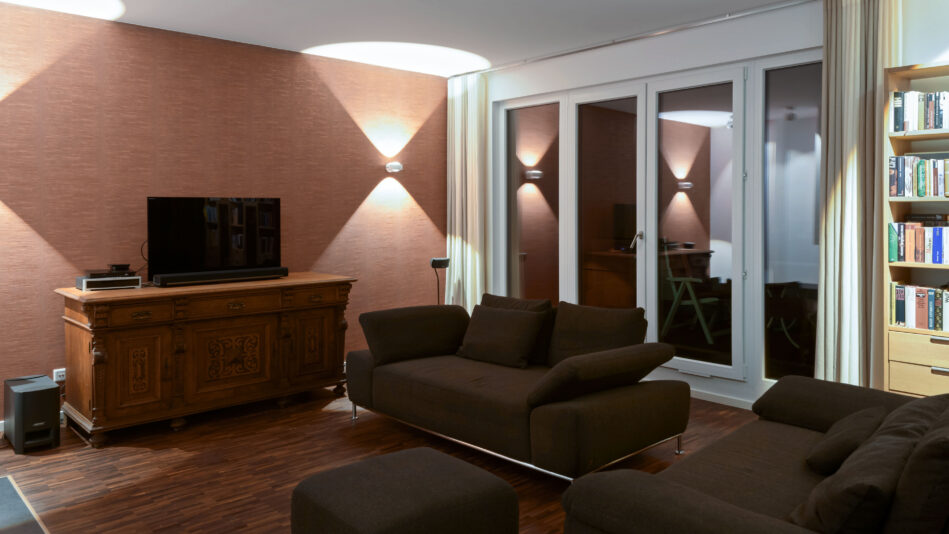 Dieses Wohnzimmer ist in warmen Rot- und Brauntönen gehalten, zwei direkt-indirekt strahlende Wandleuchten links und rechts des Fernsehers sorgen für eine stimmungsvolle Allgemeinbeleuchtung. (Foto: Prediger Lichtberater)