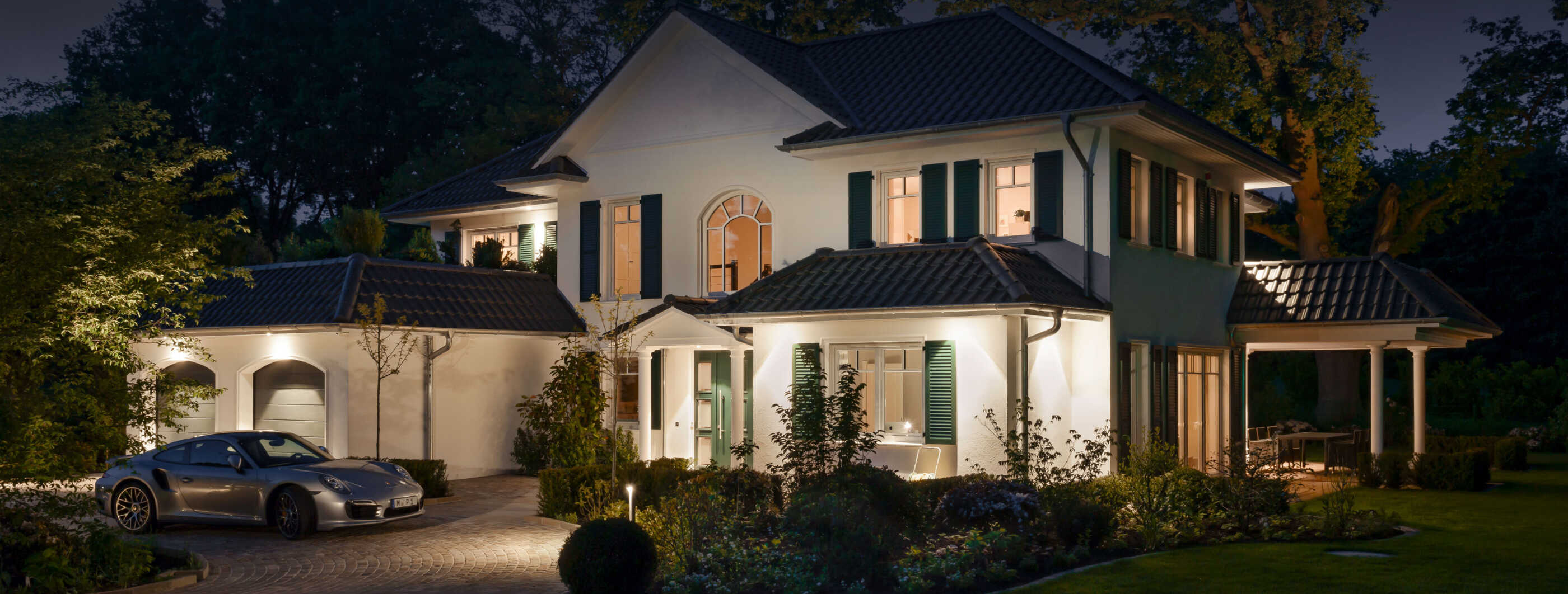 Eine luxuriöse Villa bei Nacht. In der Einfahrt steht ein Sportwagen, die Fassade wird durch Einbaudownlights punktuell beleuchtet. (Foto: Prediger Lichtberater)