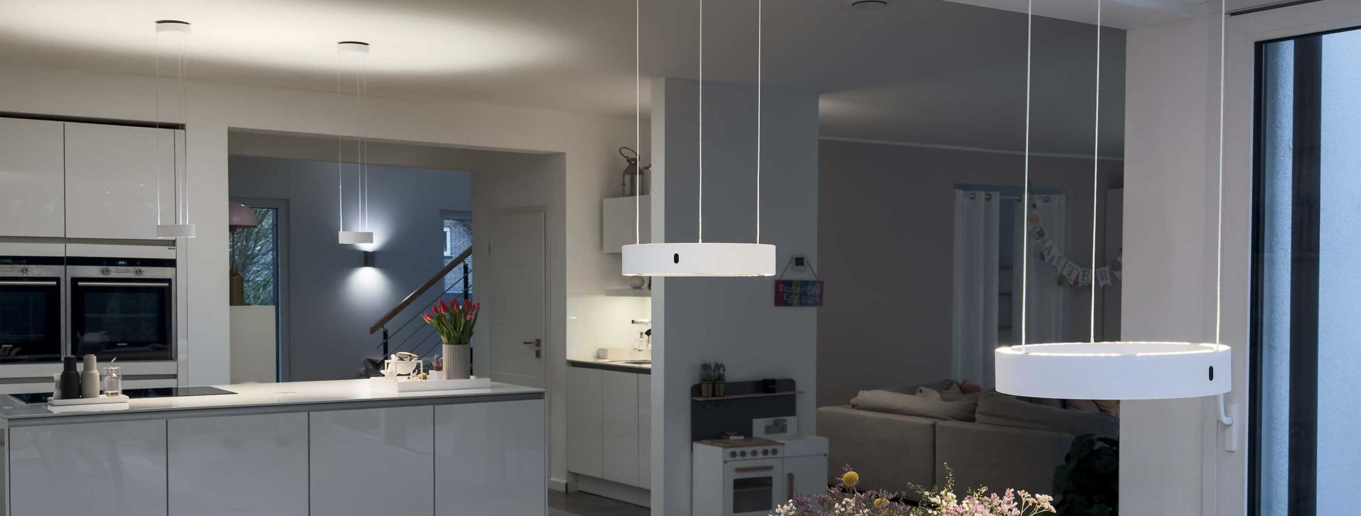 Über einem Esstisch hängen zwei weiße Pendelleuchten von CHRISTOPH (c.Space pendant), im Hintergrund ist eine offene Küche zu sehen, in der die kompakteren c.Pace pendant für gute Beleuchtung sorgen.